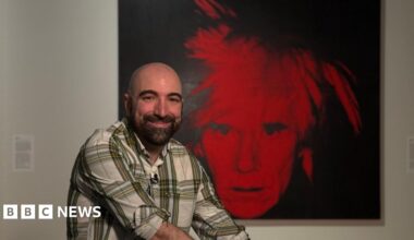 A man smiling in a gallery in front of a black and white self portrait by Andy Warhol