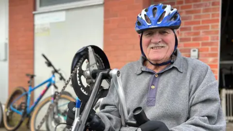 BBC Alan is wearing a helmet and gloves, adjusting the handlebars of a red adapted tricycle outside a brick building, with several bicycles parked nearby