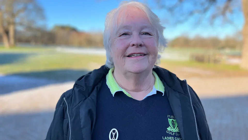 Gillean Fuller, a woman with short grey hair, wears a blue overcoat over a blue jumper and green golf shirt, and looks directly into the camera.
