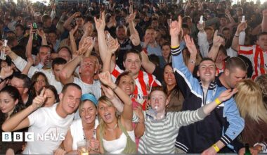 A picture taken from the stage of a crowd of people singing and dancing with arms in the air. Several are wearing red and white-striped Sunderland shirts, in one corner there is a couple kissing. A man at the front with short ginger hair is blowing a whistle, there are two young women with blonde hair cheering next to him and many people are clutching plastic drink glasses and bottles.
