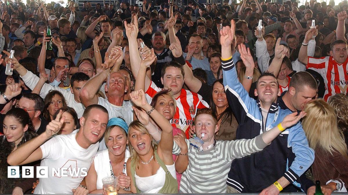 A picture taken from the stage of a crowd of people singing and dancing with arms in the air. Several are wearing red and white-striped Sunderland shirts, in one corner there is a couple kissing. A man at the front with short ginger hair is blowing a whistle, there are two young women with blonde hair cheering next to him and many people are clutching plastic drink glasses and bottles.