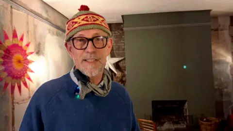 Man in hat wearing glasses and blue jumper and a scarf
