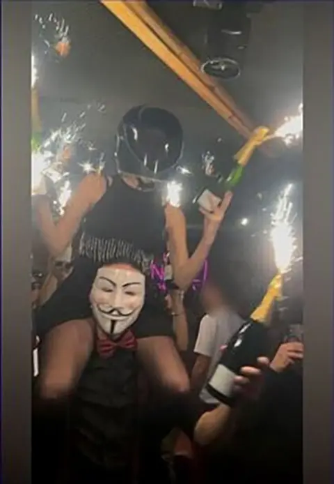 A person wearing a motorbike helmet sits on the shoulders of another person. Both of them are holding champagne bottles with sparklers attached, close to the ceiling and walking through a bar busy with people.