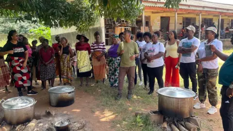 Jose Tembe/BBC  Shafee Sidat, the mayor of the Marracuene, talks to a group sheltering at a school with cooking pots set up over logs under trees 