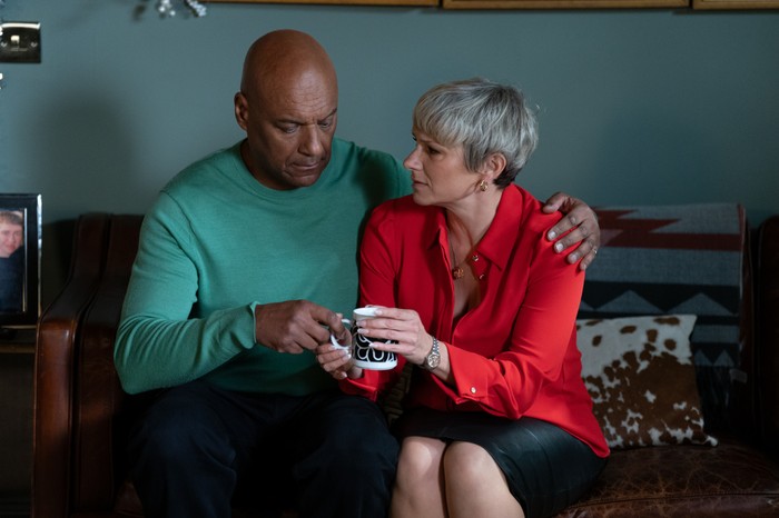 Colin Salmon as George Knight in a green jumper sat beside Laura Doddington as Nicola Mitchell in a red blouse with his arm around her shoulder for EastEnders.