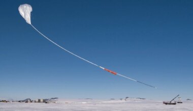 A white scientific balloon rises into the bright blue sky above snowy Antarctica. The sky and the balloon take up most of the image, with the ground only being a small white stripe at the bottom of the photo. The balloon has a long white "tail" with orange and black portions.
