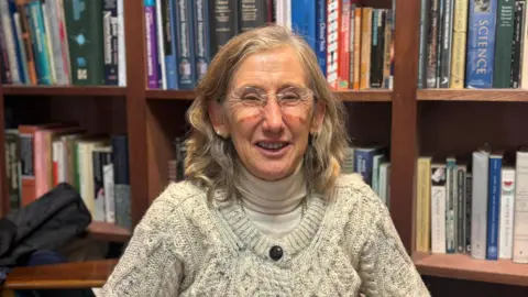 Orla Hardiman has long, fair hair. She is wearing a cream top with a white polo neck jumper underneath. She is smiling and has blue eyes. A wooden book case is behind her.