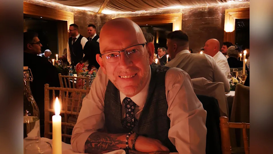 Mr Burke in a white shirt and dark waistcoat smiling at the camera with his arms crossed on the table in front of him. He Has dark glasses and is sitting down at an event, with a candle burning on the table just in front of him. 