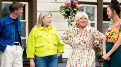 Stephen (left), Harriet, Fiona and Roxy stand on a train platform. Fiona wears a floral midi dress and has blonde short hair and a fringe, while Harriet wears a yellow short jacket. Stephen has short mousy brown hair and wears white jeans and a blue denim jacket. Roxy wears a brightly patterned blouse and smiles at the others.