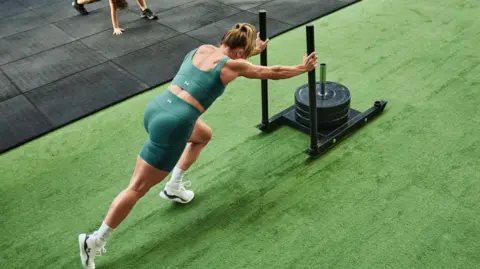 @vicswilliamson Vicky Williamson is wearing a green lycra top and shorts. She is pushing weights along Astroturf.