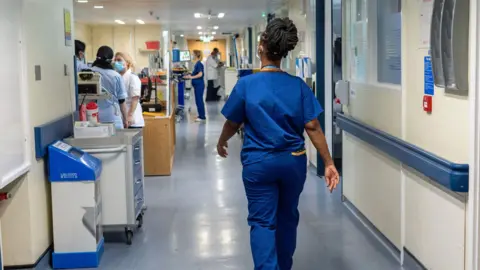 PA Media A female nurse is seen from behind walking down a hospital corridor, wearing royal blue scrubs. There are various items of hospital equipment and staff wearing masks further down the corridor.