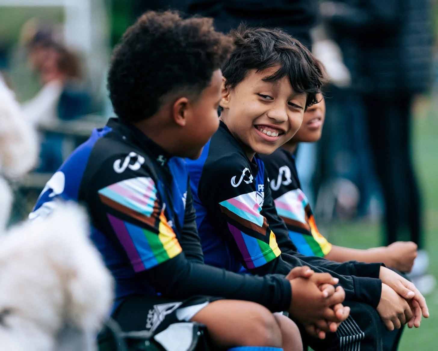 Two children smile at each other, wearing soccer jerseys emblazoned with Pride flags