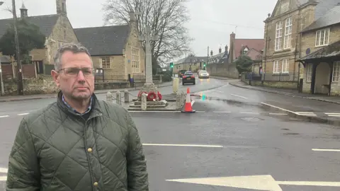 Amy Holmes/BBC A man wearing a green quilted jacket stands at the edge of a wet road in a village setting. Behind them are stone buildings, a roadside war memorial with wreaths, and traffic cones placed near the junction on a grey, rainy day.