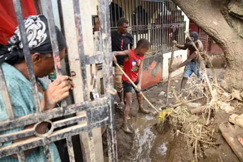 Egeder Pq Fildor / Reuters People clean up debris in front of a flooded house as a family member looks on, in Petit Goave, Haiti on 31 October 2025.