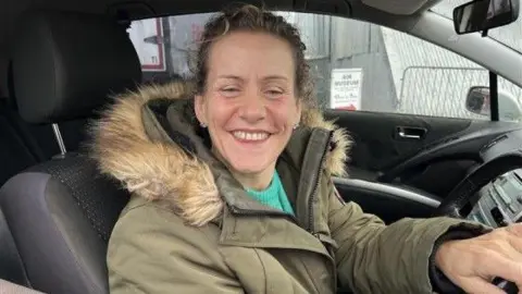 PHIL HARRISON/BBC Caroline Stone wears a parka coat as she sits in the driving seat of her car and smiles into the camera.
