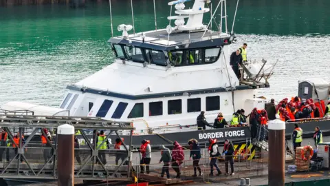 Getty Images A border force vessel carries asylum seekers into port at Dover. A line of men waering life vests can be seen disembarking the vessel and walking in a line. the boat says "Border Force" along the side