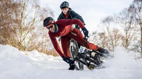 Paul Campbell A mountain biker dressed in red skids in deep snow during the Puffer. It is bright daylight in an area of woodland and the cyclist is watched by a younger competitor behind him.