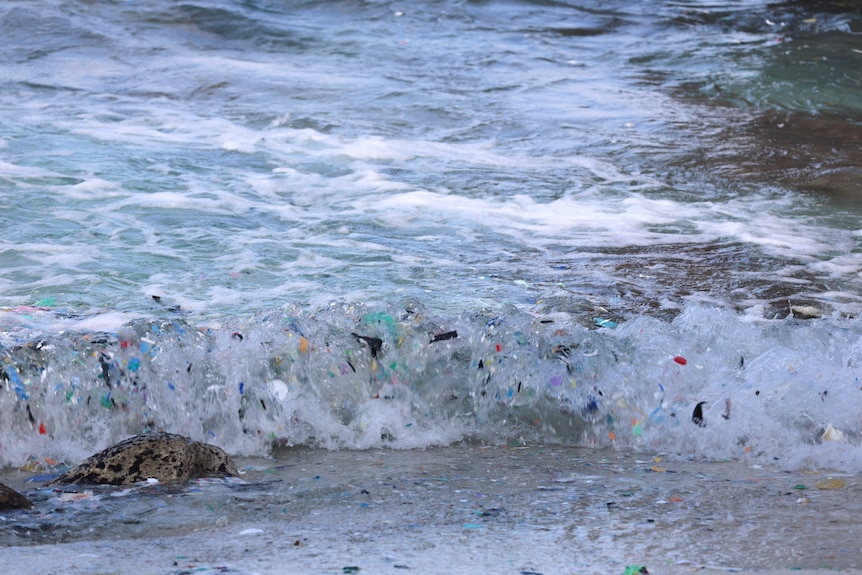 A small wave crashes with small coloured pieces of plastic in it.