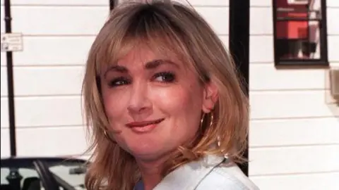 A candid shot of Caroline Aherne has her looking off to her left in front of a white building. She wears a pale denim jacket and has her hair in a blonde shoulder-length style. There is a car in the street behind her.