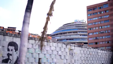 El Helicoide prison is seen in the distance with a wall in front of it that has a picture of Nicolás Maduro