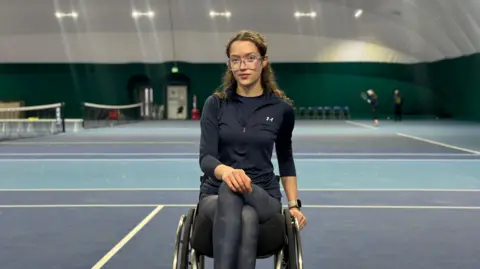 Alice Dyer has curly brown hair and is wearing clear glasses. She's on her wheelchair wearing navy blue sportswear. She's at a tennis court in the Nottingham Tennis Centre