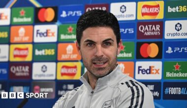 Mikel Arteta speaking at his news conference in a grey Arsenal tracksuit top, with a board of sponsor logos behind him