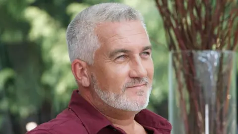 Getty Images Paul Hollywood is seen in a closeup photograph on a baking show. He is wearing a shirt and has grey hair and a beard and he is smiling. There is a vase of stems behind him from a flower arrangement.