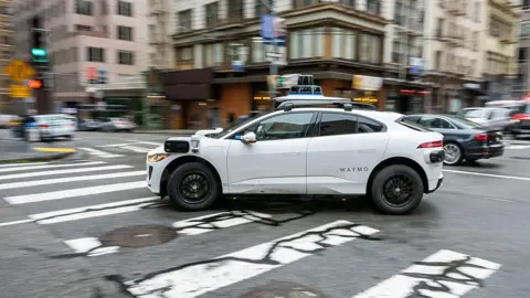 Bloomberg A white Waymo autonomous taxi driving down the road.