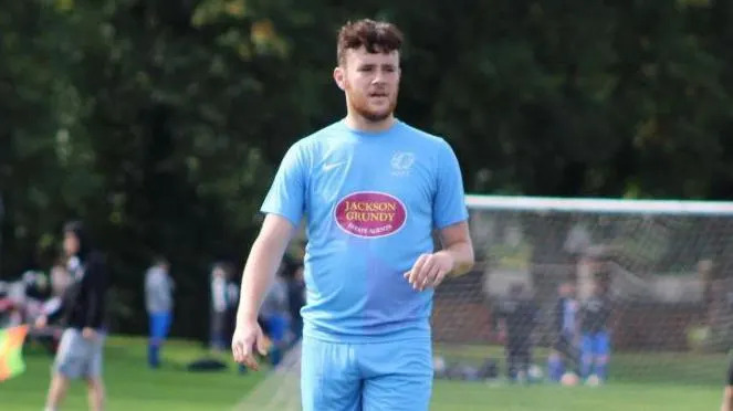 Luke Abrahams is on a football pitch during a match. He is wearing a light blue T-shirt an shorts and a football net can be seen in the blurred background.