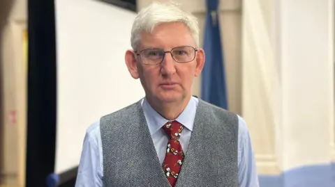 A grey haired man with glasses looks at the camera, wearing a blue shirt, grey waistcoat and red tie.