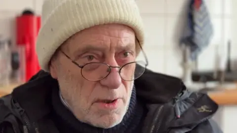 Close up shot of Reinhold wearing the wooly, cream hat he's been sleeping in at night.
