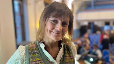BBC A woman with light brown hair, wearing a white blouse and green tank top, looks at the camera