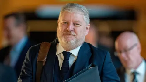 PA Media A man in a dark suit and tie carrying a leather‑strapped bag and a folder, walking indoors with several other suited figures in a softly lit corridor behind him.