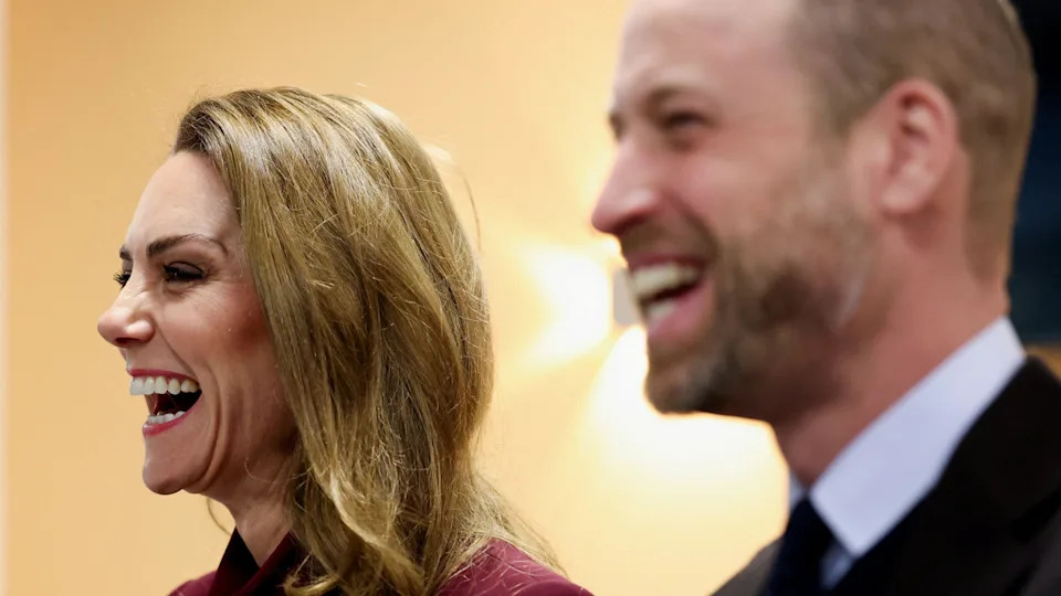 The Prince and Princess of Wales laughing in a side view. Catherine, wearing burgundy, is in focus while William, wearing a suit, is blurry.
