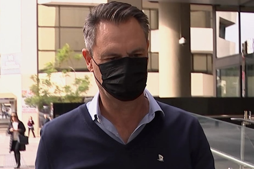 A man wears a mask, blue jumper and business shirt as he leaves court