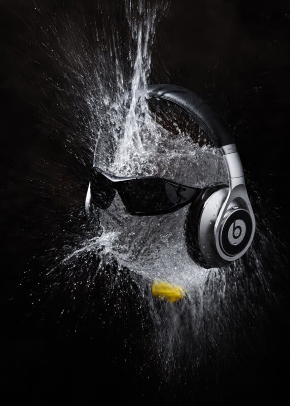 A pair of sunglasses and Beats headphones being struck by a splash of water, captured in mid-air against a black background, with droplets and waves spreading outward.