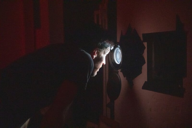 A person leans forward in a dark room, illuminated by the glow of a round light or lens in front of their face, creating dramatic shadows and a mysterious atmosphere.