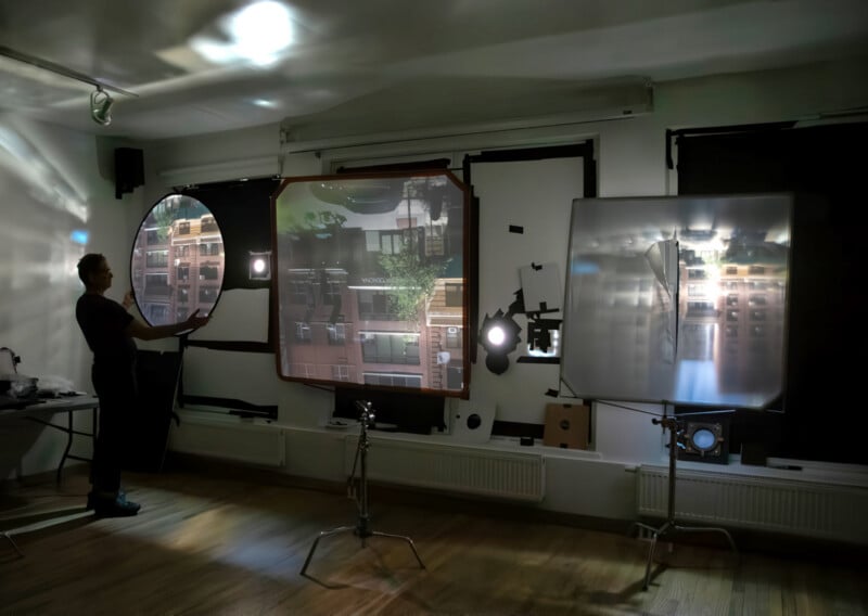 A person stands in a room with darkened windows, holding a circular reflector. Projected images of buildings appear on large screens, with light sources and equipment arranged around the wooden floor.