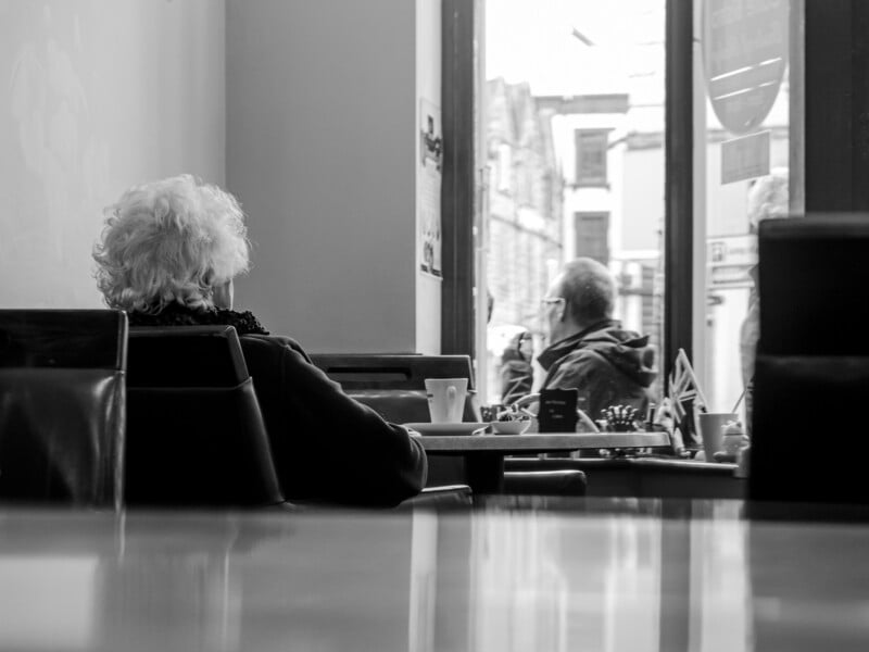 An elderly person with white hair sits alone at a table in a café, facing the window. Outside, another person is seen walking past. The scene is in black and white, creating a quiet, contemplative mood.