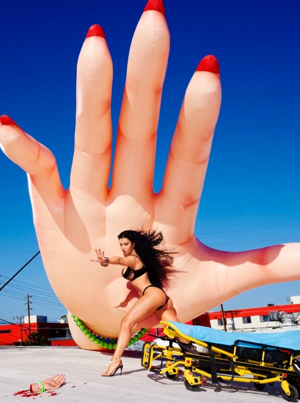 A woman in black lingerie and heels poses dramatically in front of a giant inflatable hand on a rooftop. Nearby, a stretcher and a prop severed hand are visible under a bright blue sky.