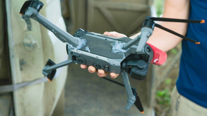 A person holding a damaged black drone with broken and bent propellers, missing parts, and visible repair attempts with glue. The background is blurred, showing a door and some outdoor elements.
