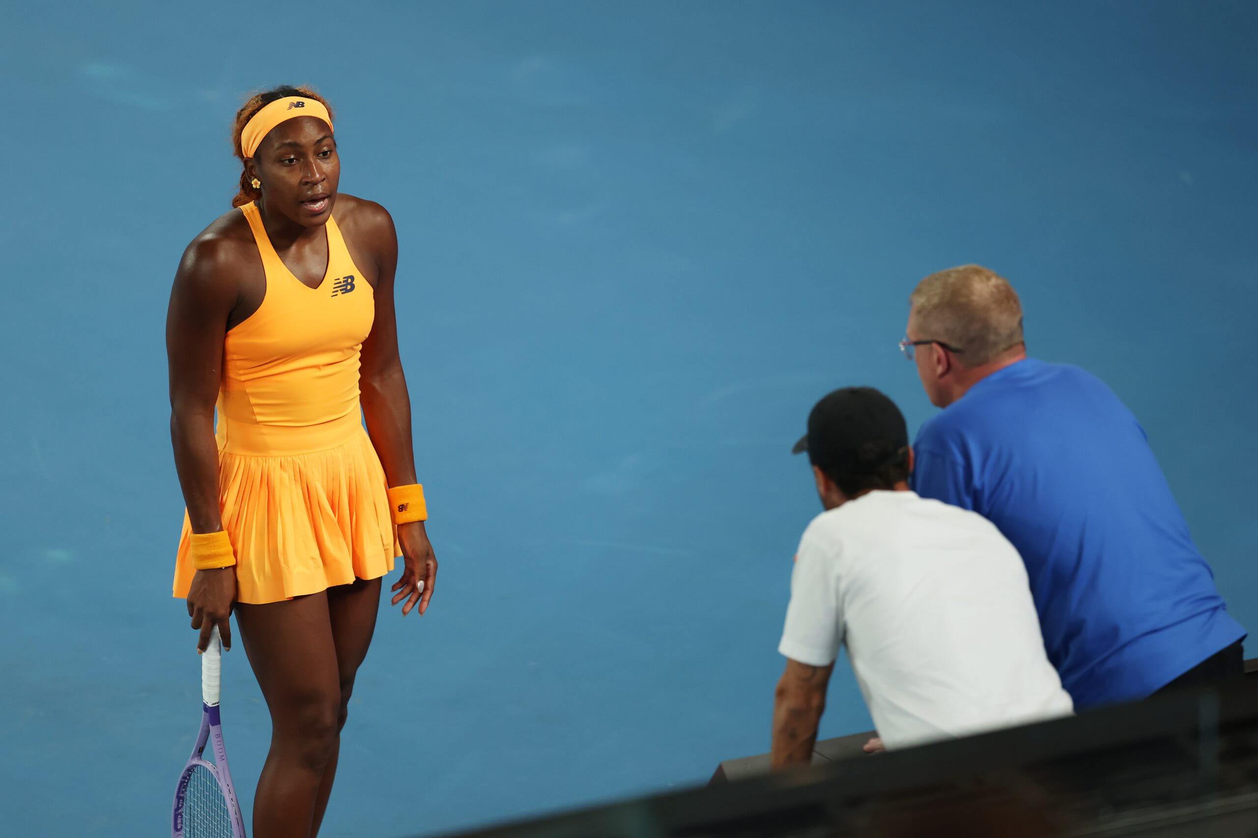 Coco Gauff stands wearing an orange tennis kit talking to two men in white and blue t-shirts.