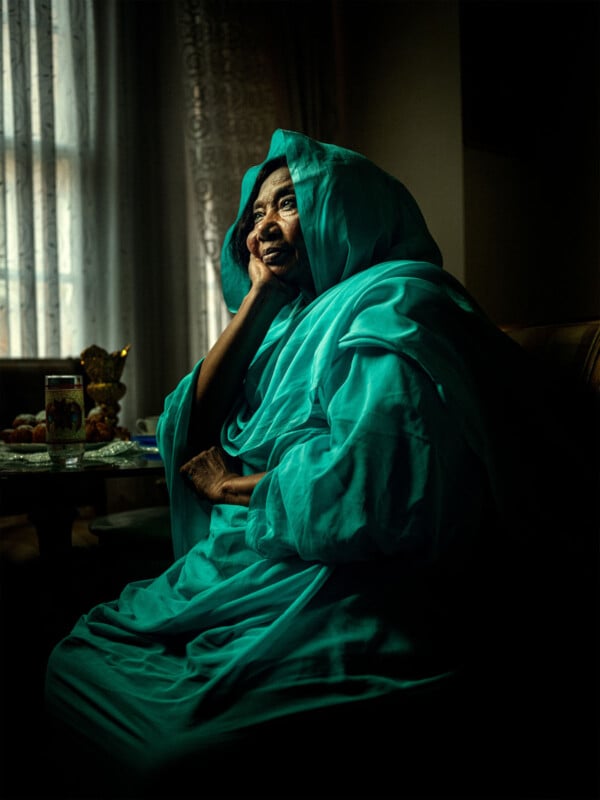 An elderly woman in a turquoise robe sits by a window, resting her head on her hand and gazing thoughtfully into the distance. Soft light from the window illuminates her face and robes.