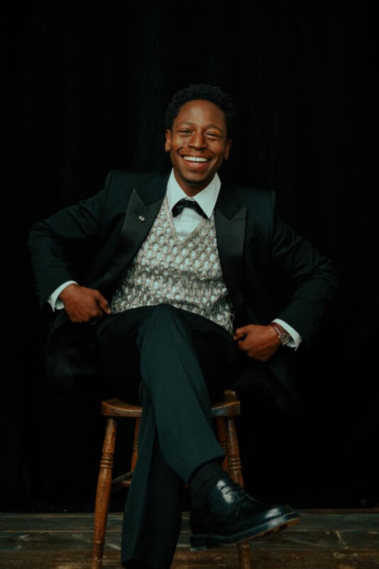A well-dressed man in a tuxedo and patterned vest sits on a wooden stool, smiling broadly against a dark background.