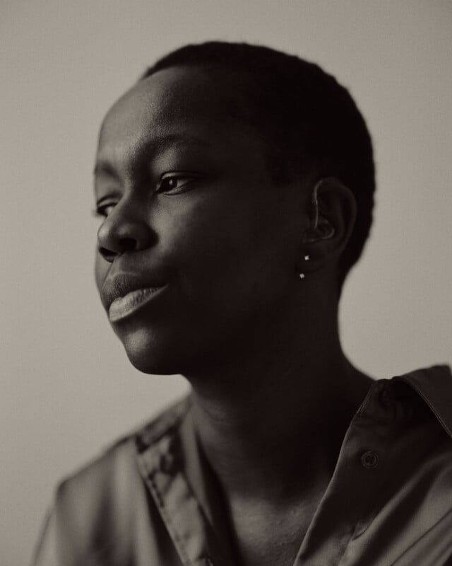 A black and white portrait of a person with short hair, wearing a collared shirt and multiple earrings, gazing thoughtfully to the side against a plain background.