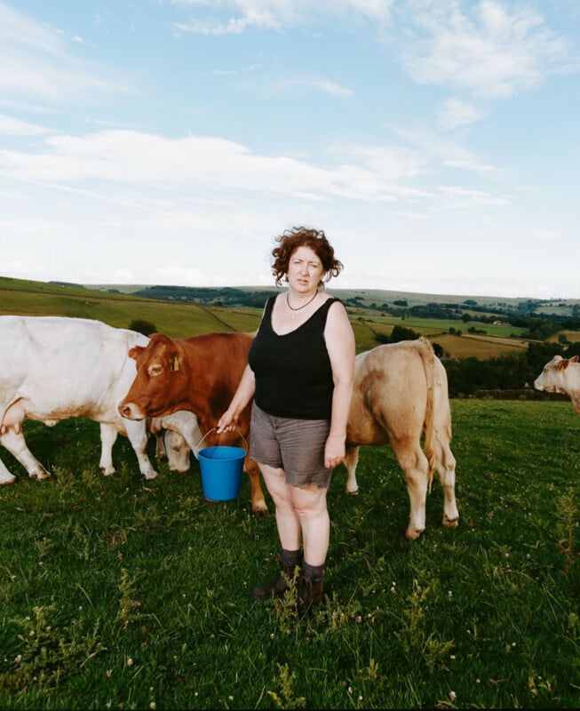 A woman in a black tank top and shorts stands on a grassy hillside holding a blue bucket, surrounded by cows grazing under a partly cloudy sky with rolling hills in the background.