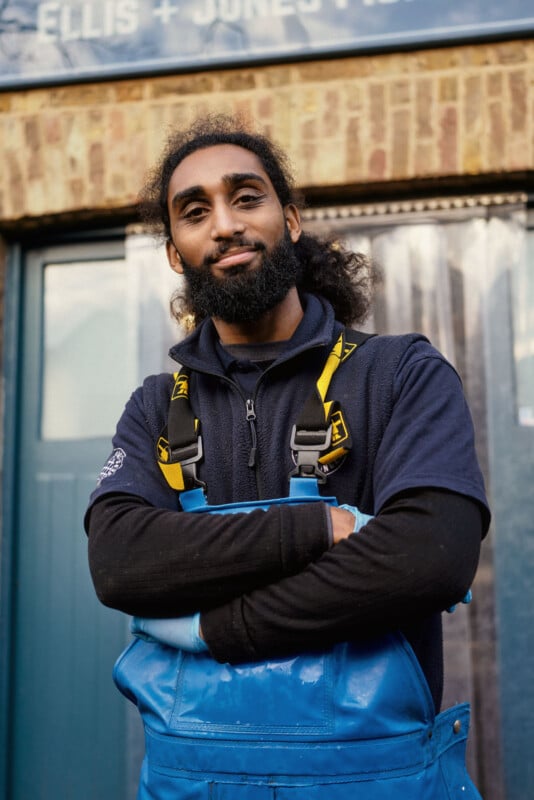 A man with a beard, wearing a navy shirt and bright blue overalls with yellow straps, stands with his arms crossed and smiles confidently in front of a brick building.