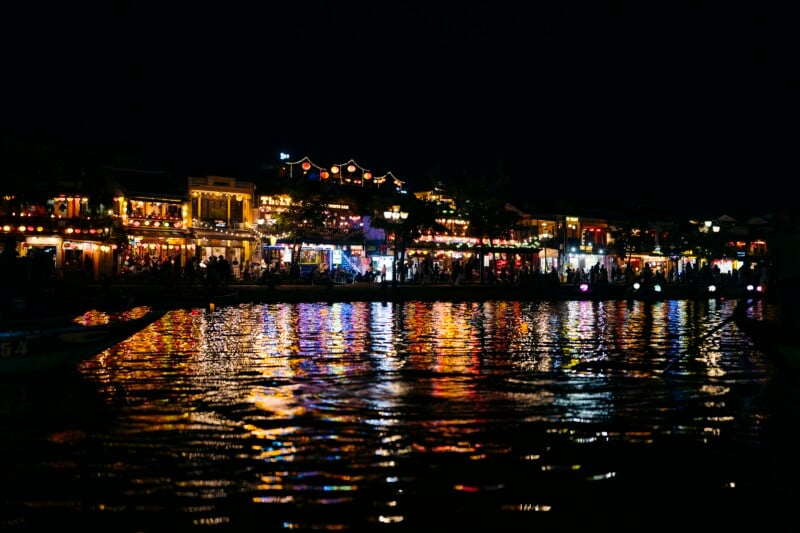 A nighttime riverside scene with colorful lights from buildings reflecting on the water, creating a vibrant and festive atmosphere. People are visible along the waterfront, adding to the lively ambiance.
