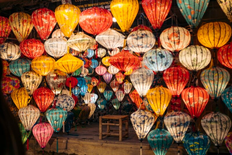 Colorful paper lanterns of various shapes and patterns hang densely together, creating a vibrant and festive display. A small wooden stool sits in the background amidst the lanterns.