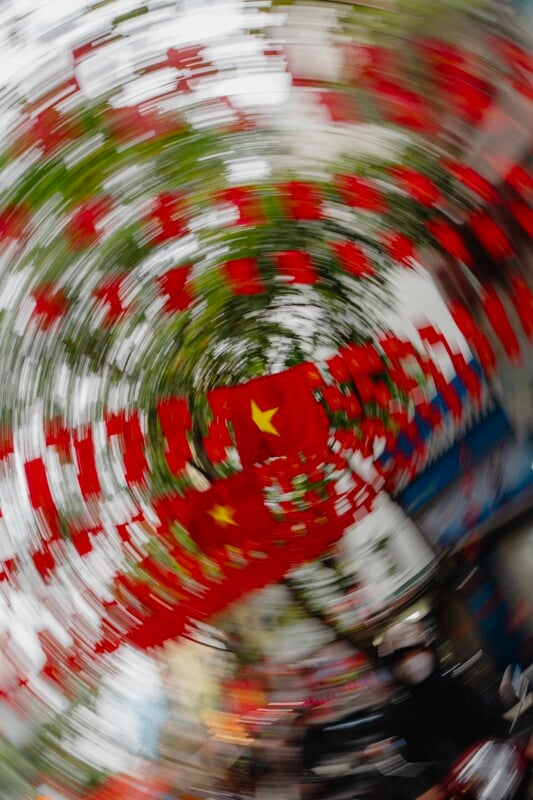 A swirling, motion-blurred image of many red flags with yellow stars, likely Vietnamese flags, hanging above a street lined with trees and buildings. The spiral effect creates a dynamic, dizzying visual.